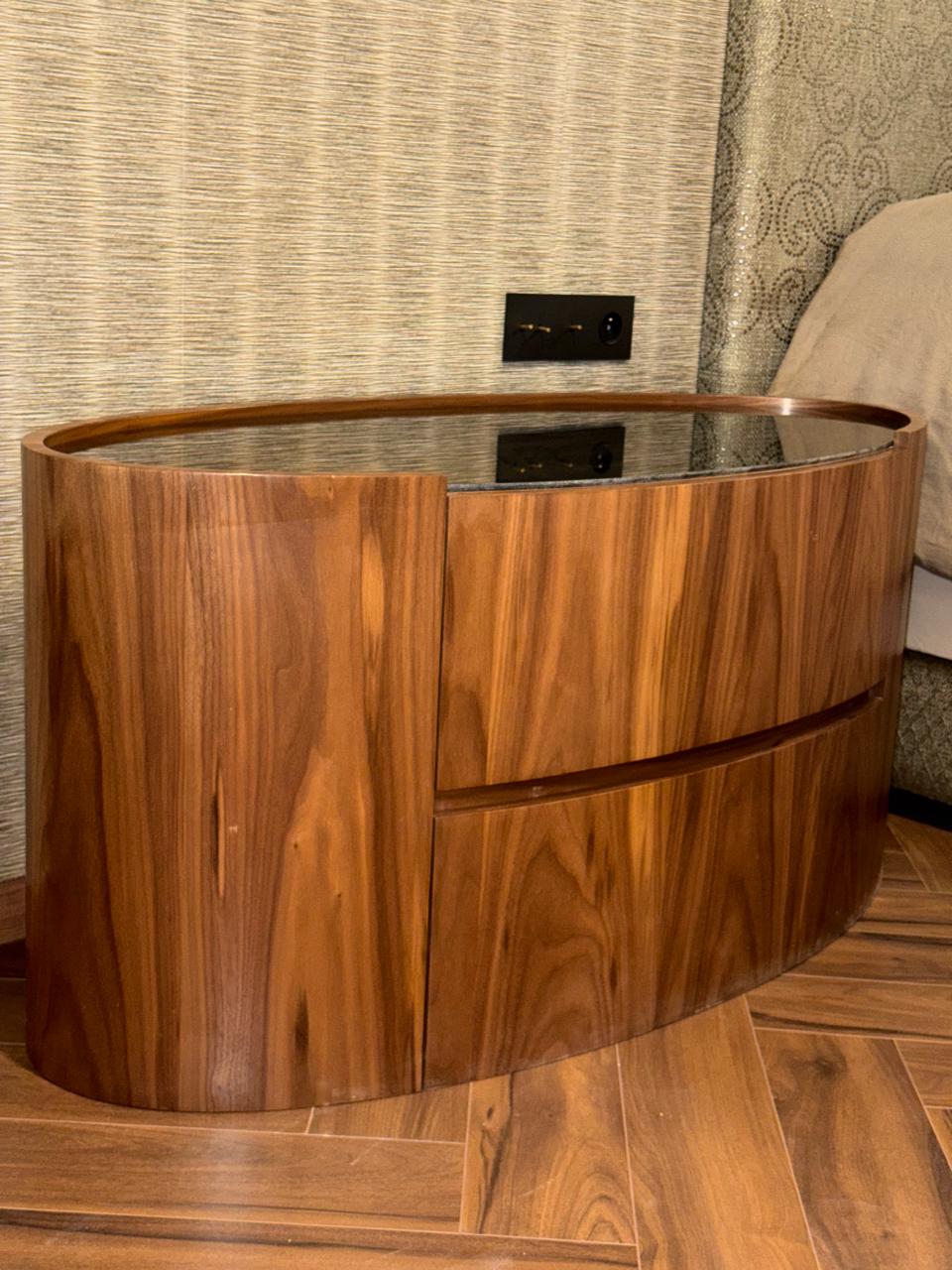 Table de chevet ovale en noyer à deux tiroirs, plateau en verre fumé, au pied d'un lit