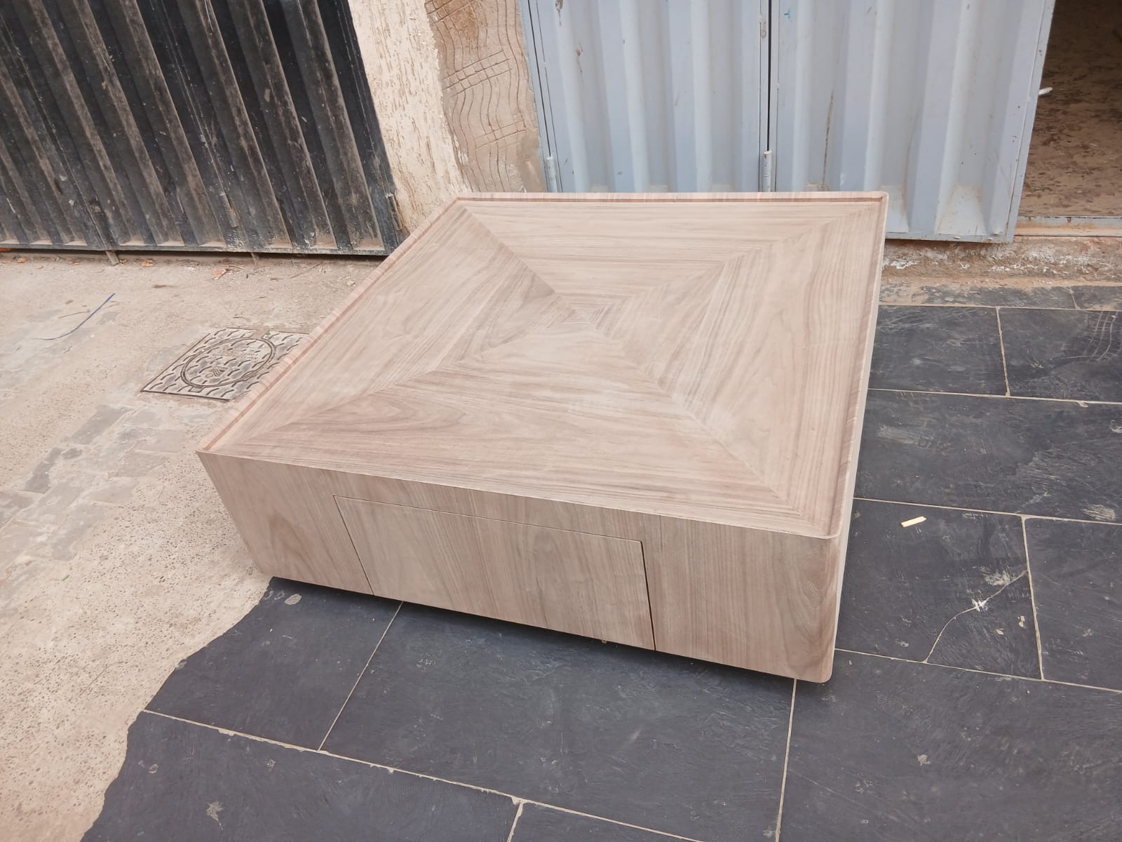 Table basse carrée en bois clair, plateau en pointe de diamant, devant la porte de l'atelier